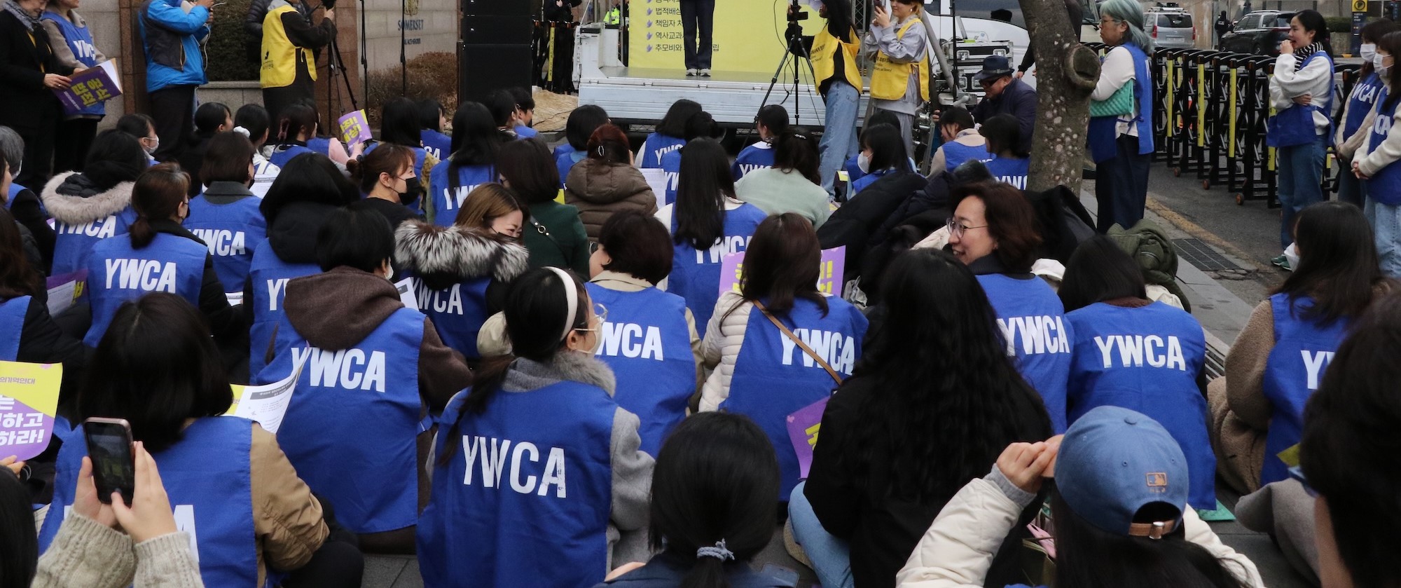 2024년 2월 14일, 한국YWCA연합회가 주관한 제1635차 수요시위 모습이다. &lsquo;YWCA&rsquo;가 적힌 파란 조끼를 입은 참가자들이 손팻말을 들고 무대를 향해 바닥에 앉아 있다.