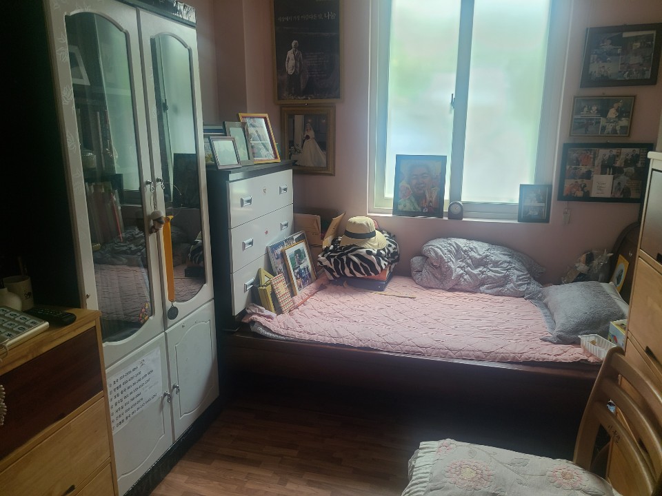 침대 주변을 가까이에서 찍은 모습이다. 유리문 장식장과 흰 서랍장 위에 액자들이 놓여 있고, 창가에 초상화 액자가, 침대 위에 밀짚모자와 이불이 놓여 있다.